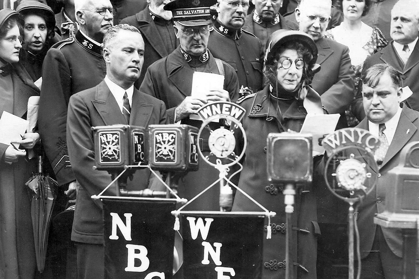 General-elect Evangeline Booth is welcomed back to New York on the steps of City Hall, with Mayor Fiorello La Guardia standing to her right, Sept. 14, 1934. Salvation Army officers and supporters stand alongside, with NBC, WNEW, and WNYC microphones capturing the event.