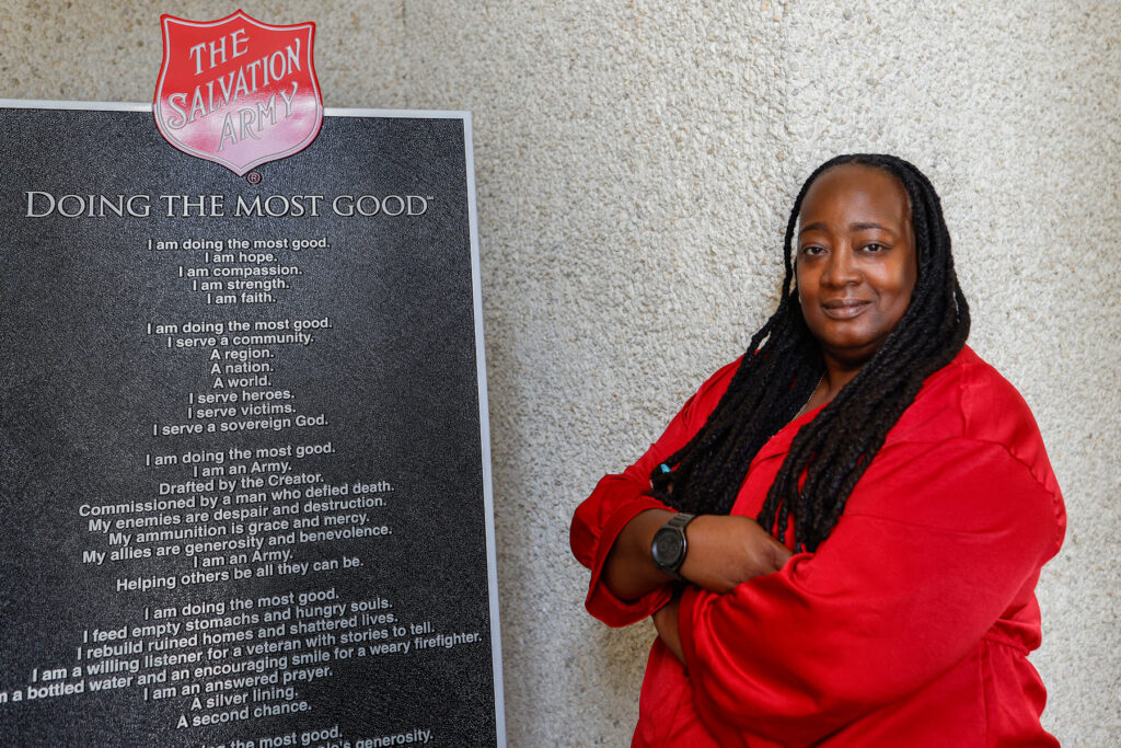 Kali Lewis, dressed in a red shirt, stands confidently with arms crossed beside a Salvation Army plaque that reads ‘Doing the Most Good,’ showcasing her dedication to ministry and community service.