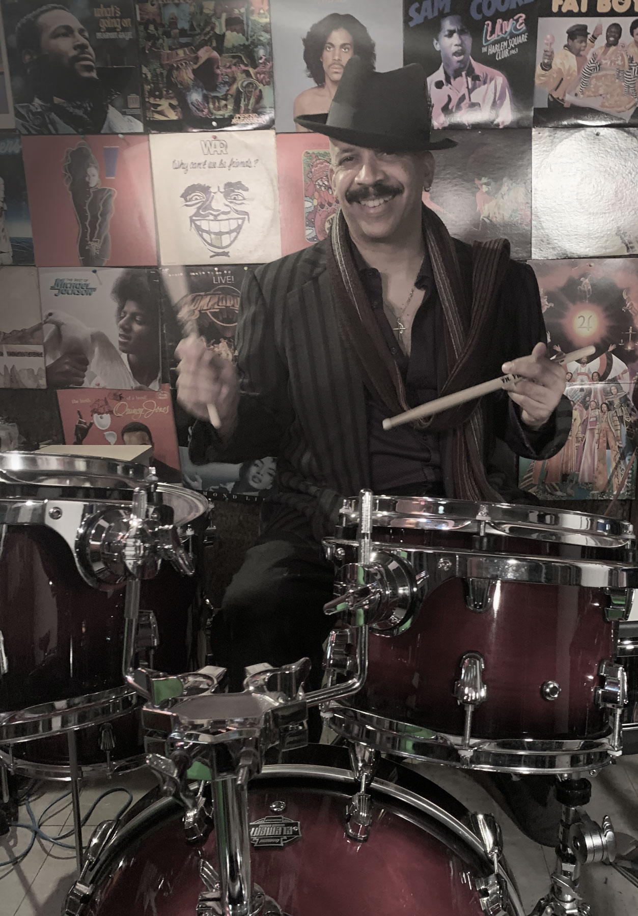 Bill Jolly, wearing a stylish black hat and striped suit, smiles while seated behind a drum set, holding drumsticks. He is surrounded by a vibrant wall of iconic album covers, reflecting his passion for music and his career as a celebrated musician.