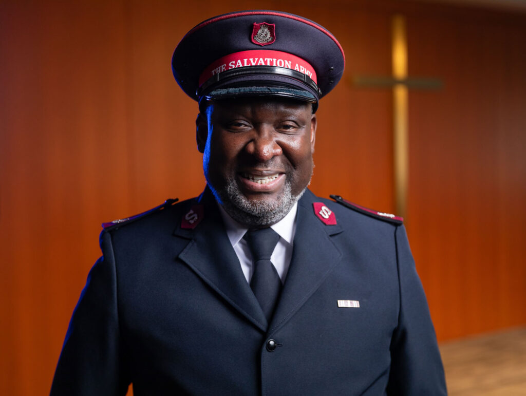 Major Elijah Kahn, dressed in his Salvation Army uniform and cap, smiles warmly in front of a softly lit background featuring a wooden cross, symbolizing his faith and leadership within the organization.
