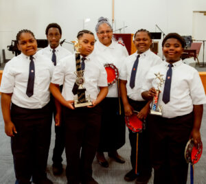 Major Cynthia Foster stands proudly with a group of young boys from the timbrel team, all dressed in formal Salvation Army attire. The boys hold their timbrels and display trophies, celebrating their achievements and growth through teamwork, dedication, and faith.
