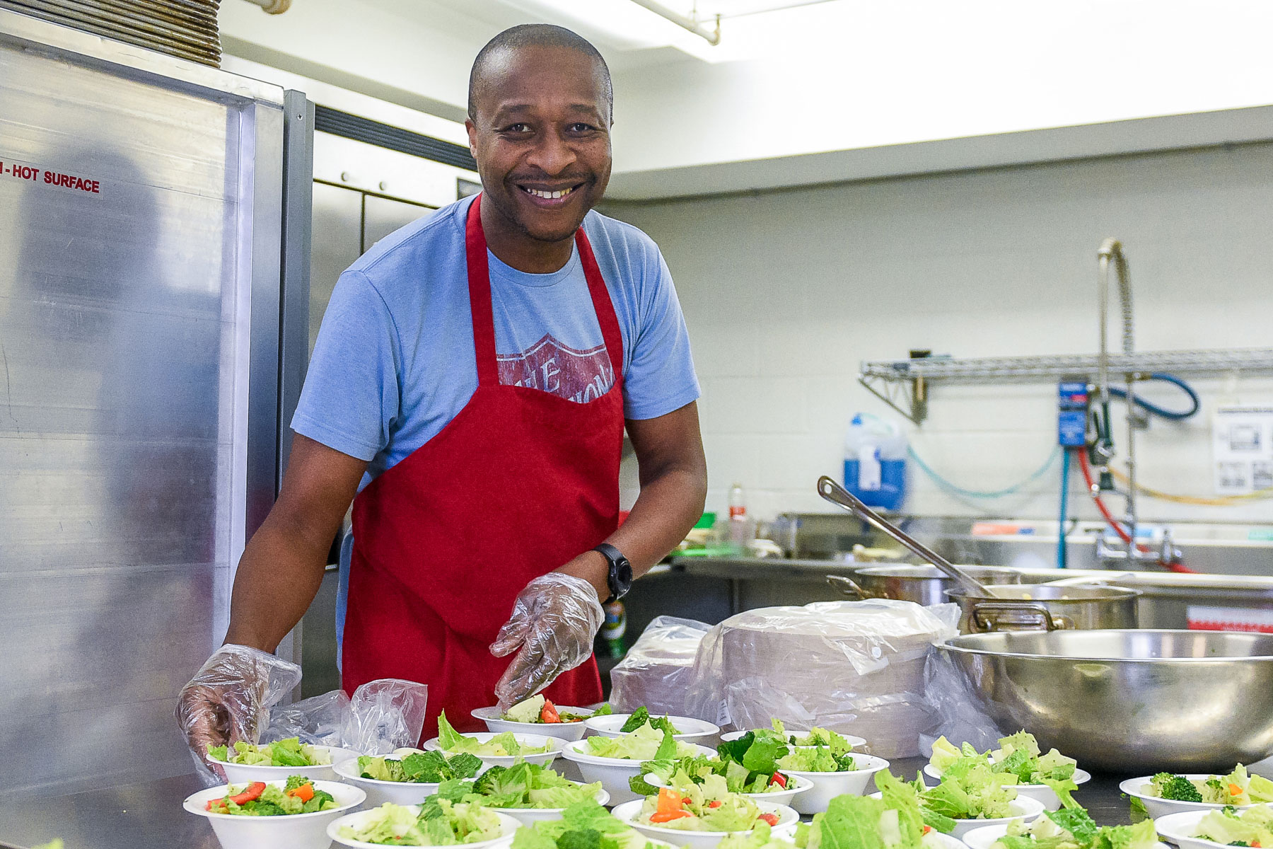 A smiling volunteer wearing a red apron and gloves prepares fresh salad bowls in a kitchen, showcasing dedication and care in serving meals to the community.