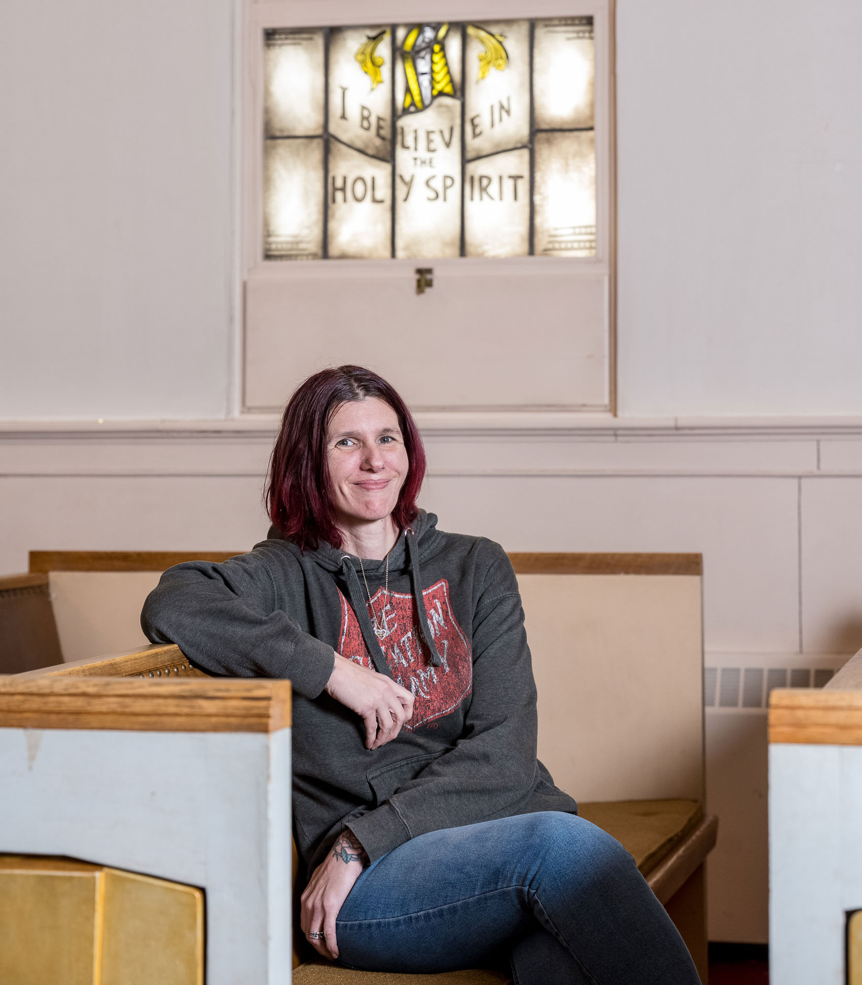 A woman with shoulder-length red hair sits in a church pew, smiling warmly. She wears a dark Salvation Army hoodie with a red shield logo and blue jeans. Behind her, a stained-glass window displays the words ‘I BELIEVE IN THE HOLY SPIRIT.’ The church interior features wooden pews and cream-colored walls. Photo by J. Fiereck Photography.