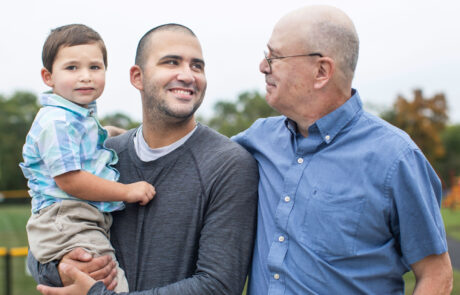 A young father, a grandfather, and a little boy stand closely together in a park. The father holds the child in his arms while the grandfather, dressed in a blue shirt, looks warmly at his son. Their smiles and affectionate postures capture the bond between three generations.