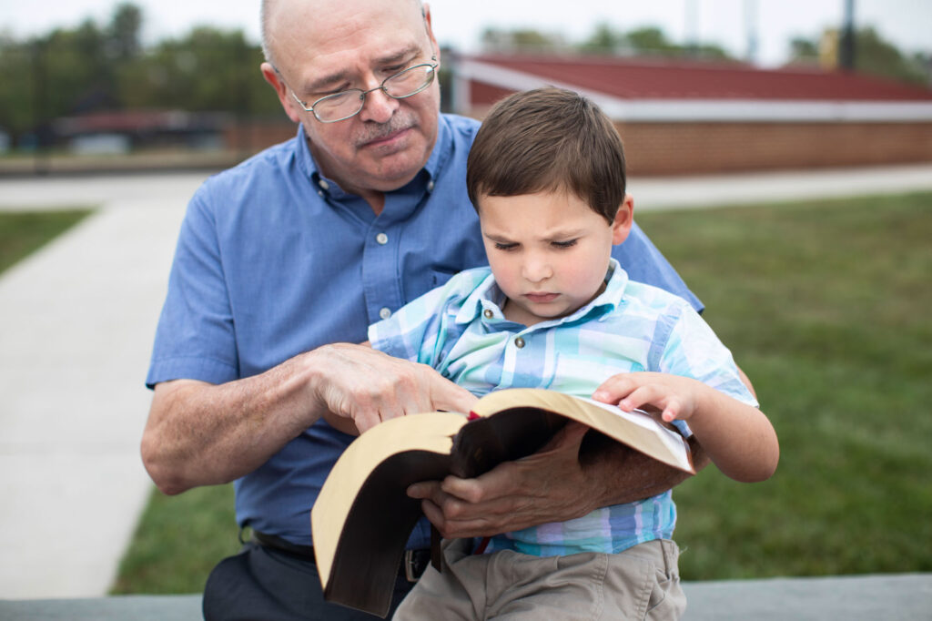 An elderly man wearing glasses and a blue button-up shirt sits outdoors on a bench, reading a bible with a young boy on his lap. The boy, dressed in a plaid shirt, looks intently at the pages while the man points to a passage. They share a moment of learning and connection in a peaceful outdoor setting.