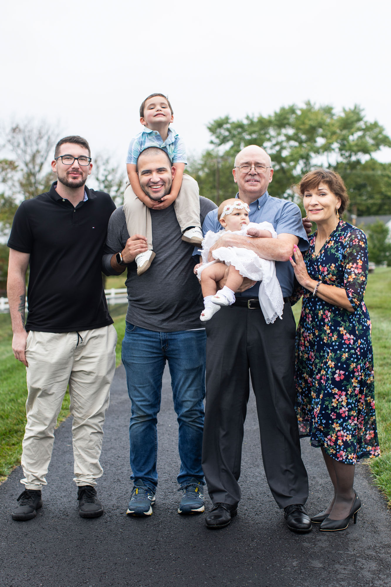 A multigenerational family of the author stands together outdoors on a paved path, smiling for the camera. An older man in a blue button-up shirt holds a baby dressed in white, while a young boy sits on a man’s shoulders. A woman in a floral dress and another man in a black polo complete the happy group, capturing a moment of togetherness.