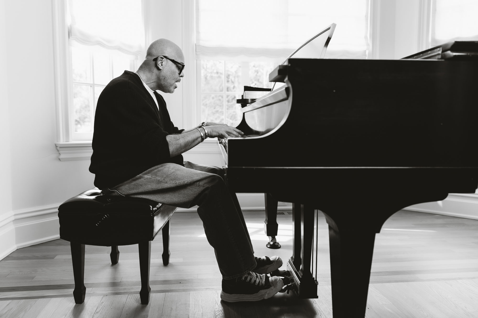 Israel Houghton plays a grand piano in a sunlit room, seated on a piano bench with his hands on the keys and eyes focused. The black-and-white photo captures him mid-performance, wearing glasses, a sweater, and jeans.