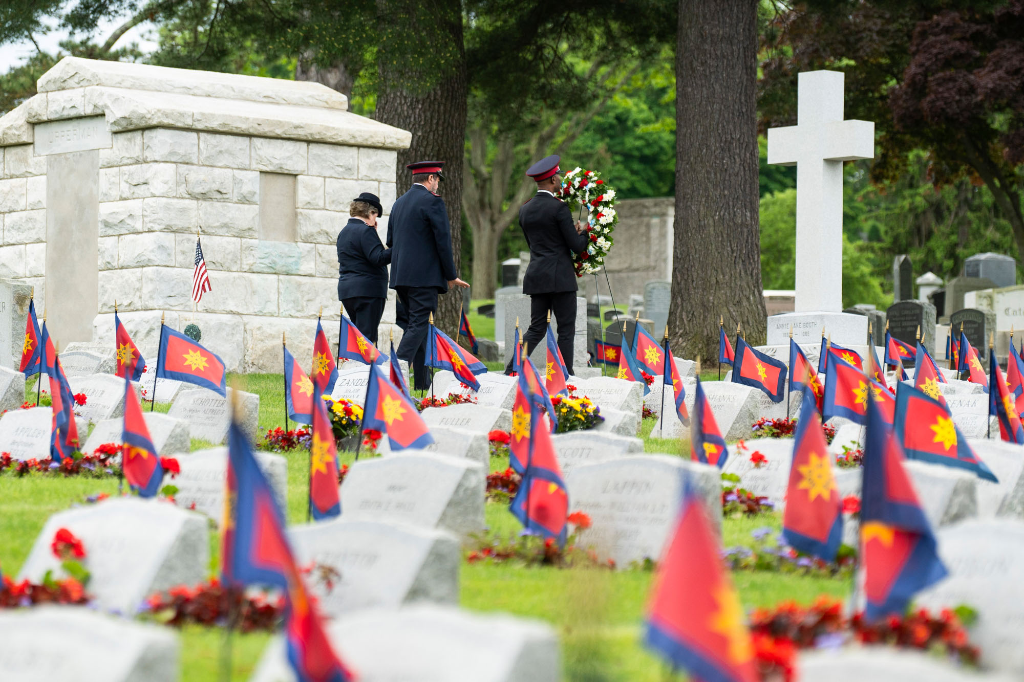 Three Salvation Army officers participate in a wreath-laying ceremony at Kensico Cemetery, surrounded by rows of headstones adorned with Salvation Army flags and red flowers. A white cross and stone mausoleum are visible in the background, set among tall trees and gravestones honoring past officers. Photo by Antonio Di Caterina.
