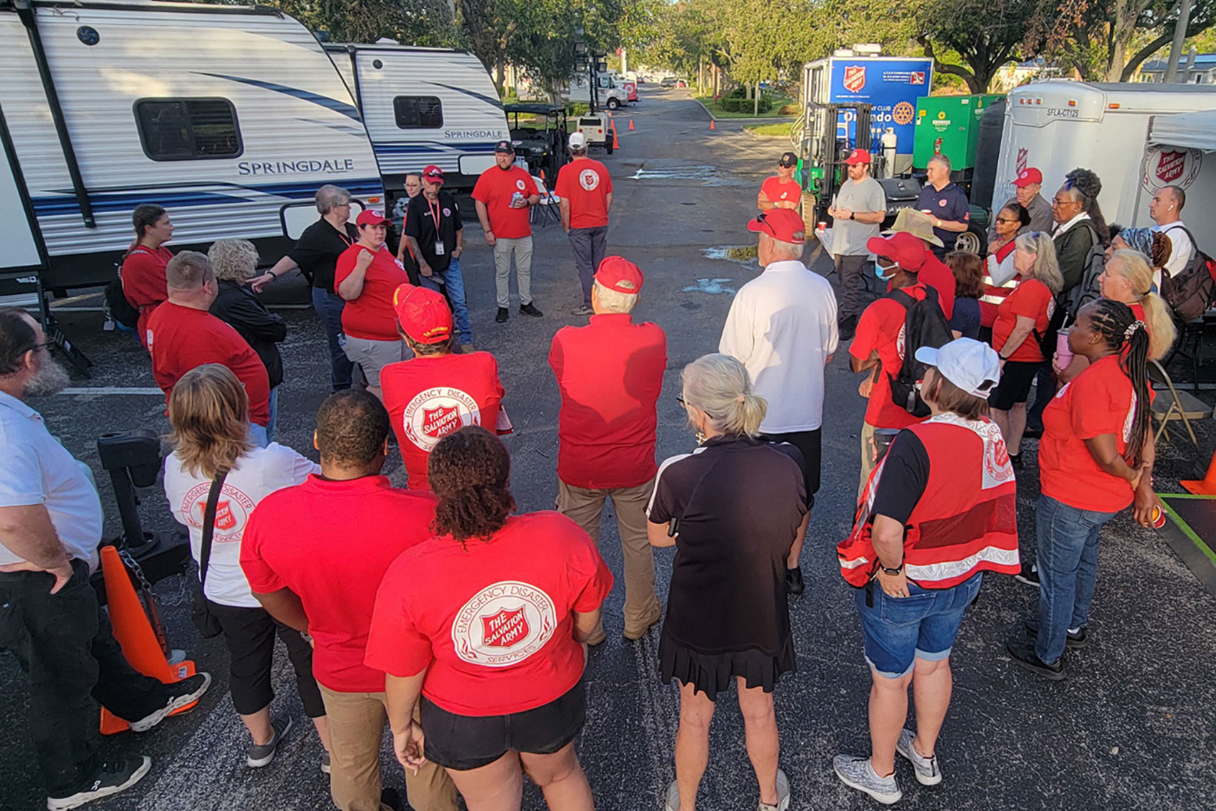 Salvation Army Emergency Disaster Services team members and volunteers stand in a circle for a morning briefing in a parking lot filled with trailers and response vehicles. The gathering takes place at the Salvation Army Social Services Center in Clearwater, Florida, following Hurricane Milton in October 2024. Most participants wear red EDS shirts or vests, symbolizing their active service in disaster relief efforts.