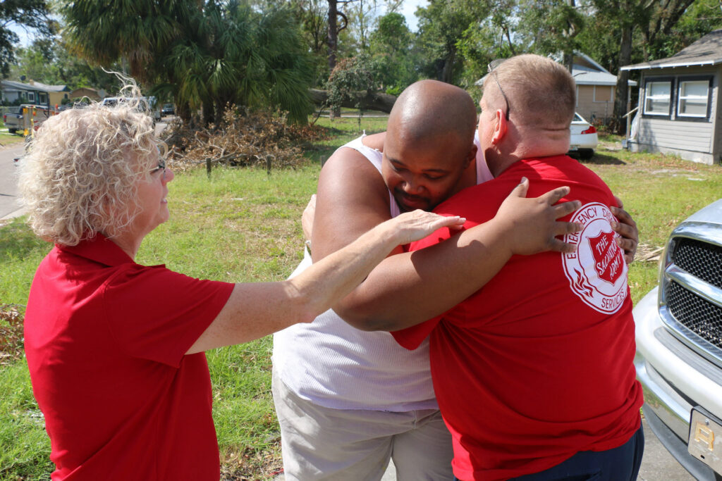 Major Candy Fritsch and Captain Edward McMillen of The Salvation Army Northeast Ohio Division embrace and comfort a man named Shelby outside his home, where debris and ruined belongings are visible. The moment reflects the emotional and spiritual care provided through Emergency Disaster Services in the aftermath of Hurricane Milton.