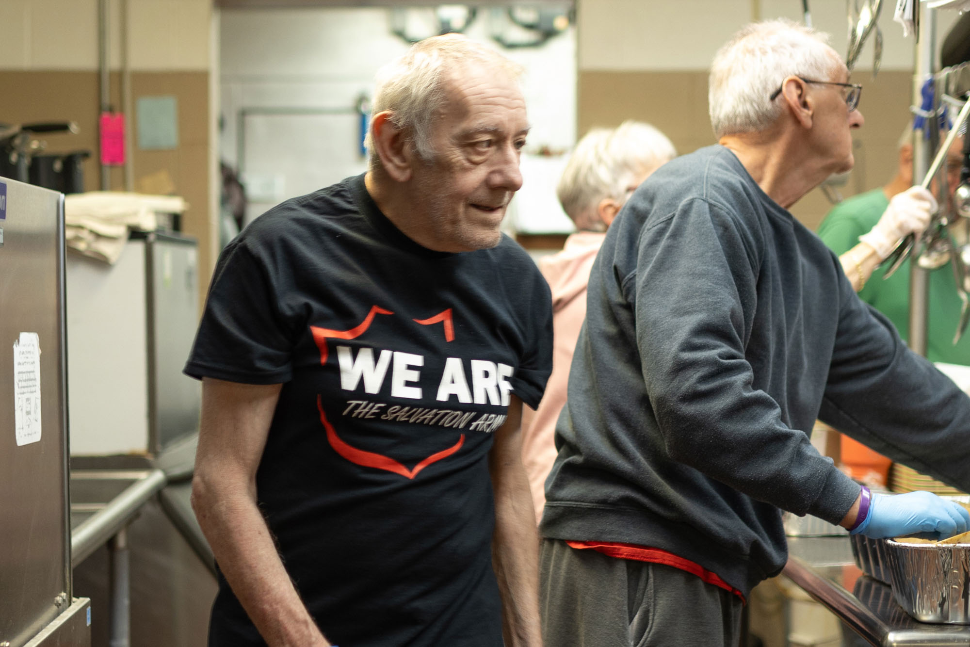 An elderly man wearing a “We Are The Salvation Army” T-shirt volunteers in a busy kitchen, helping prepare meals alongside other volunteers.
