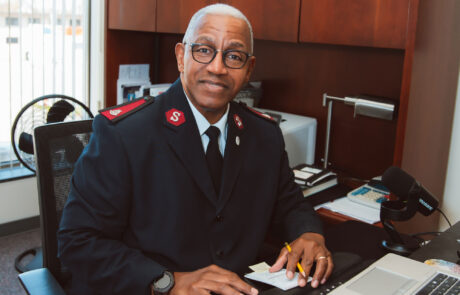 Major Charles F. Roberts sits at his office desk, smiling at the camera while holding a pencil and notepad. Dressed in Salvation Army uniform, he is surrounded by office items including a laptop, microphone, desk lamp, and fan. Photo by Emily Kae Photography.