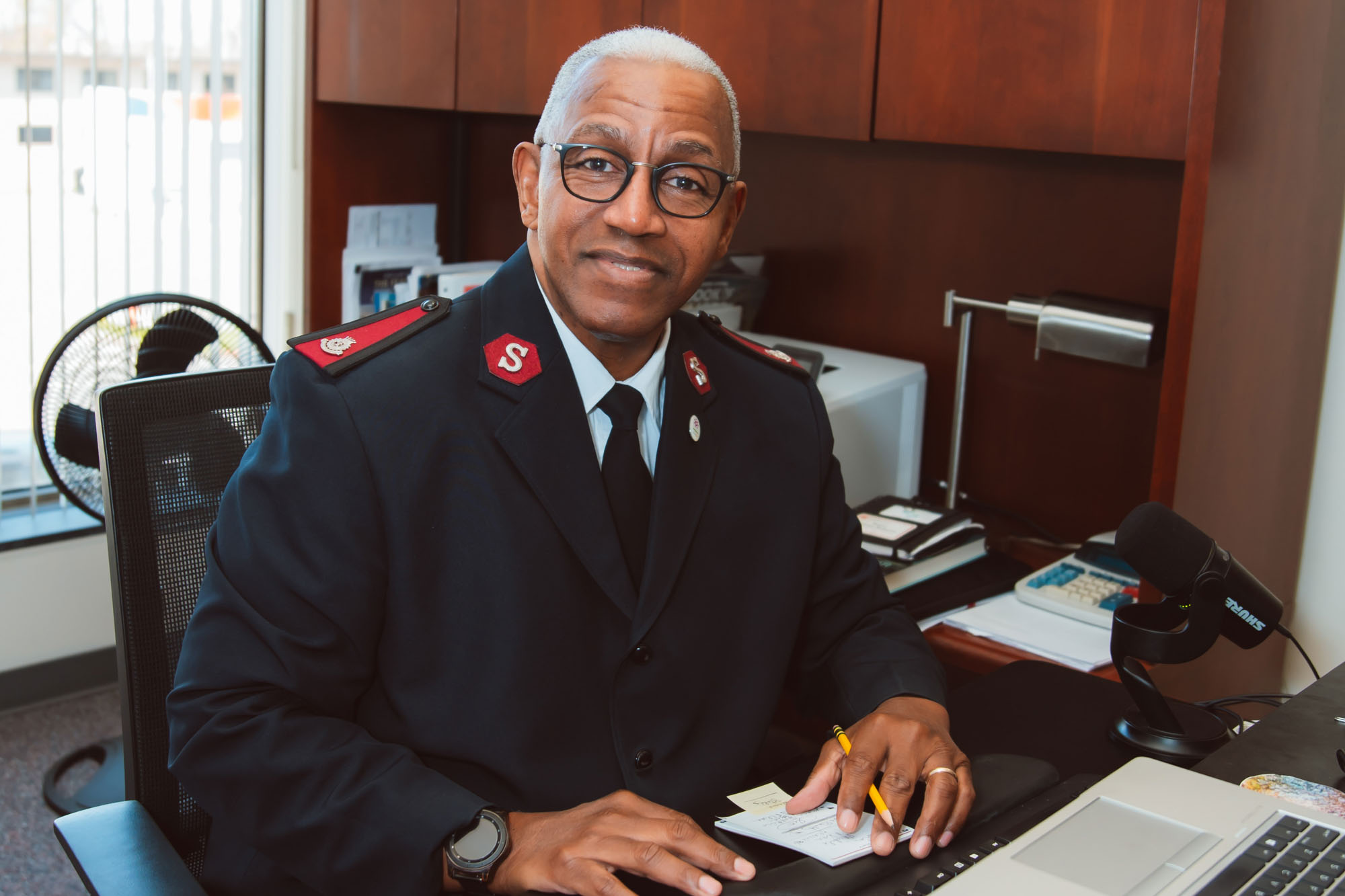 Major Charles F. Roberts sits at his office desk, smiling at the camera while holding a pencil and notepad. Dressed in Salvation Army uniform, he is surrounded by office items including a laptop, microphone, desk lamp, and fan. Photo by Emily Kae Photography.